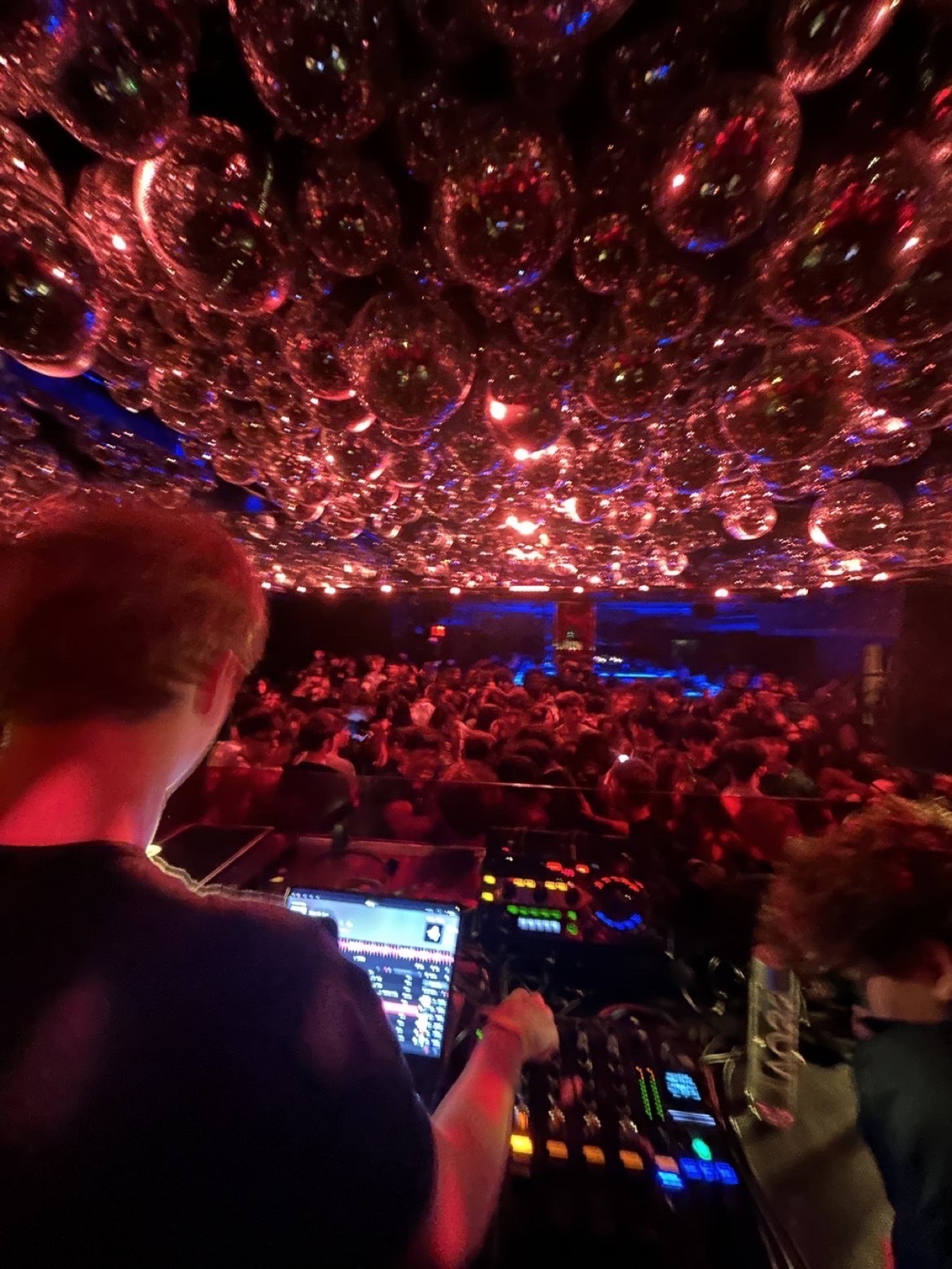 A DJ booth looking out over a packed CJL.Productions room beneath a ceiling of mirrored bubbles lit red and blue.